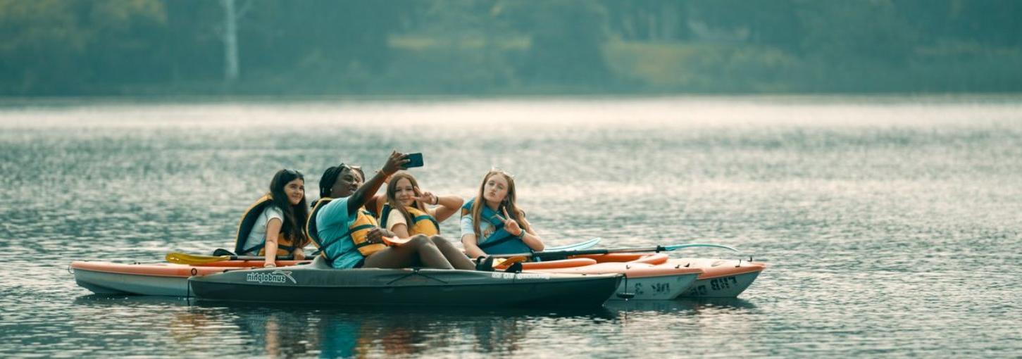 Five young girls sit on three kayaks on the water and take a selfie together.